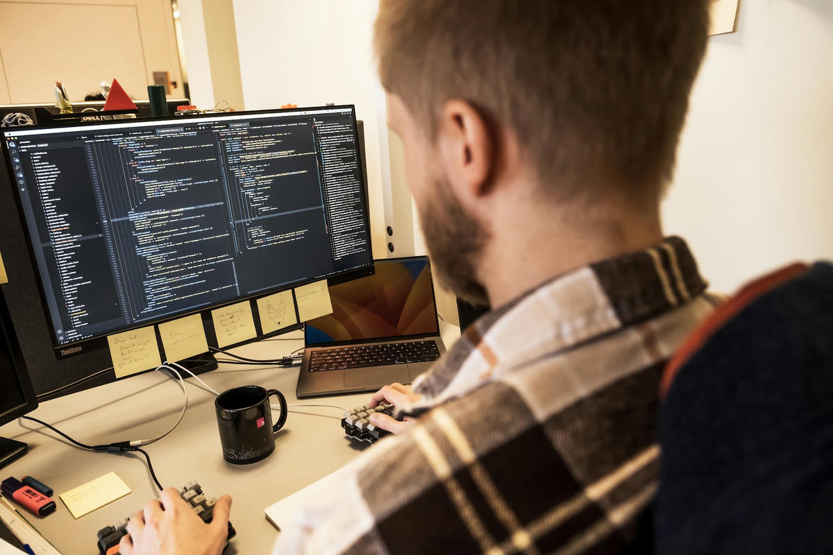 Webbutvecklare på TwoFour som arbetar vid en dator med kod på skärmen. Skrivbordet är fyllt med post-it-lappar, en kaffemugg och utvecklingsverktyg. Webbutvecklare på TwoFour som arbetar vid en dator med kod på skärmen. Skrivbordet är fyllt med post-it-lappar, en kaffemugg och utvecklingsverktyg.
