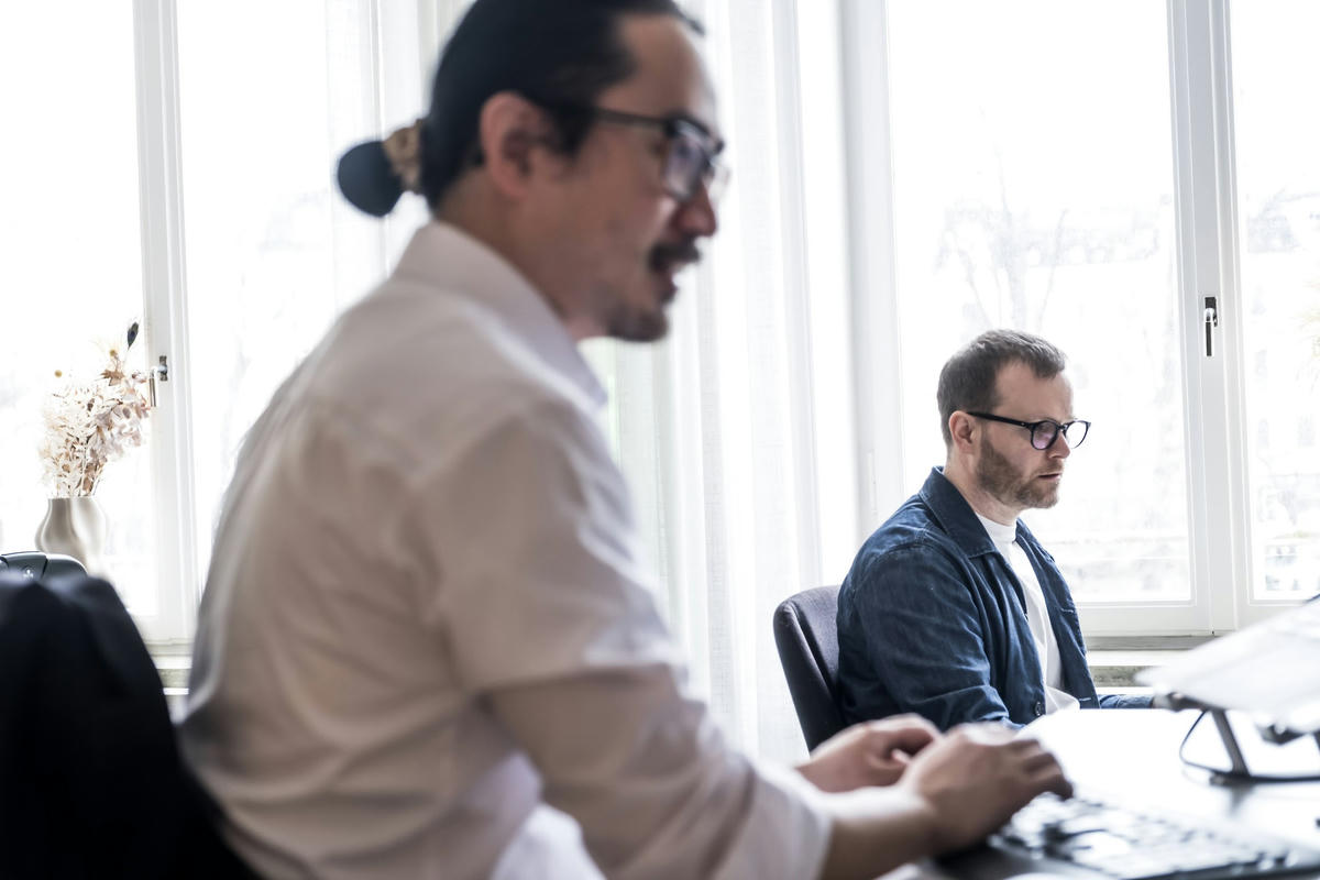 Person i vit skjorta sitter vid ett skrivbord och arbetar på sin dator, medan en annan person sitter bredvid och fokuserar på sin skärm. Båda arbetar på TwoFour i ett ljusfyllt kontorsrum.